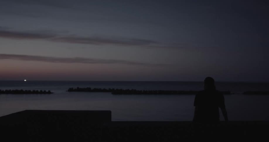 A silhouetted man sits on a concrete pier at twilight looking out over a calm ocean toward a dark breakwater and a faint orange glow on the horizon under a deepening blue sky