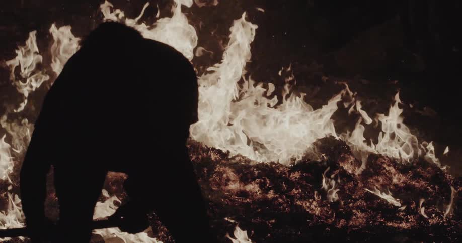 Silhouette of a man with a tool at the edge of a fire at night
