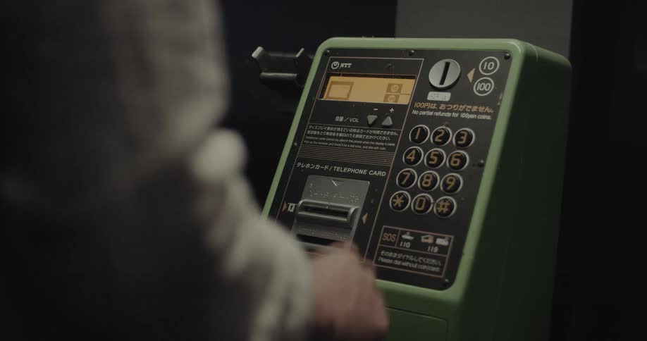 A green japanese ntt payphone with a lit display and keypad is shown dimly lit as a hand interacts with it
