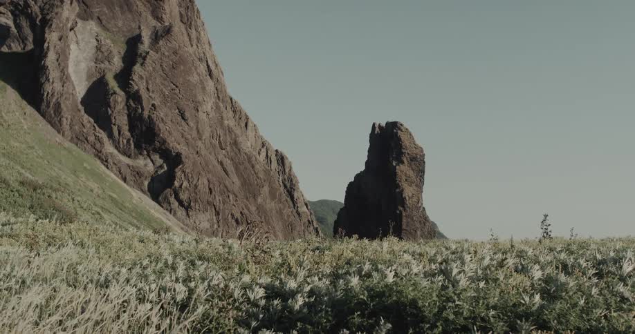 Narrow rock formation surrounded by dense vegetation and cliff to the left under clear sky