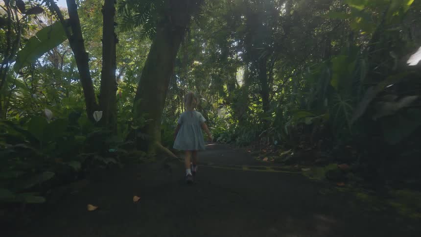 Child in blue dress and bow walking along trail in tropical garden surrounded by lush greenery