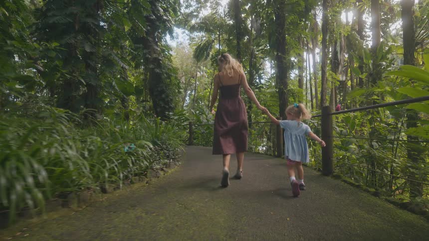 A mother and daughter holding hands as they walk and run along a forest path at midday moving excitedly beneath the bright light filtering through the trees