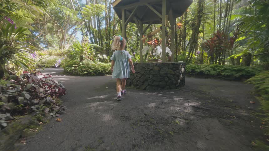 Little girl walks on a path through a tropical garden in the forest as she stops to look inside a well