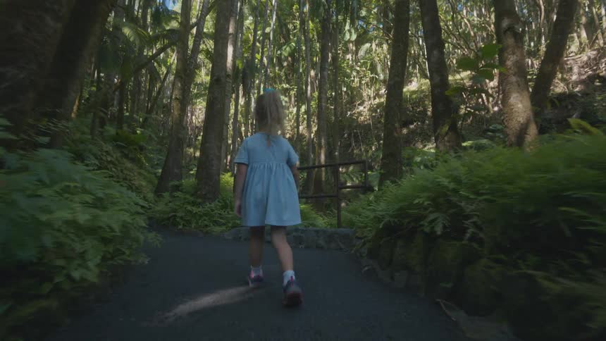 Child with blue bow walking along paved trail in lush forest under soft sunlight