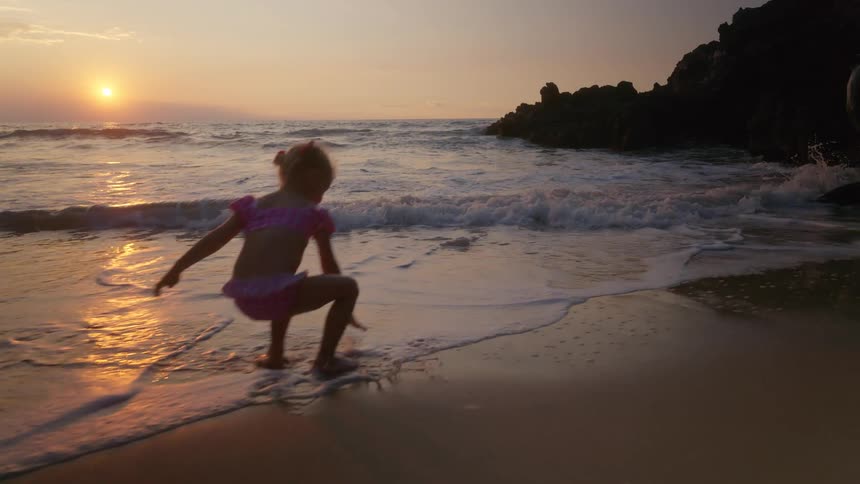 A child runs barefoot along a sunlit beach as waves crash and the sun sets behind the horizon