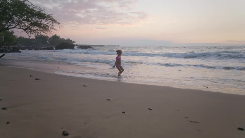Child in pink outfit playing at beach shore during sunset with colorful sky and gentle waves