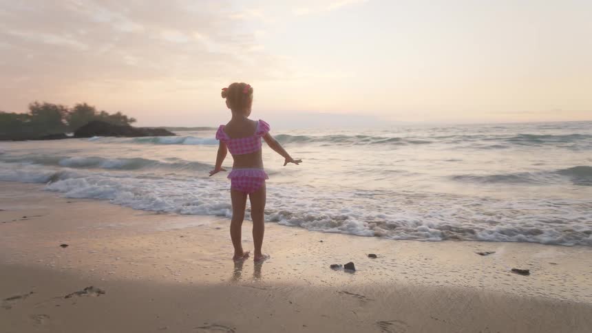 Child in swimwear standing at the shoreline with gentle waves and cliffs in the background