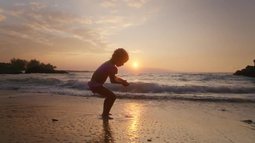 Child crouched at oceans edge during sunset with colorful sky and gentle waves around