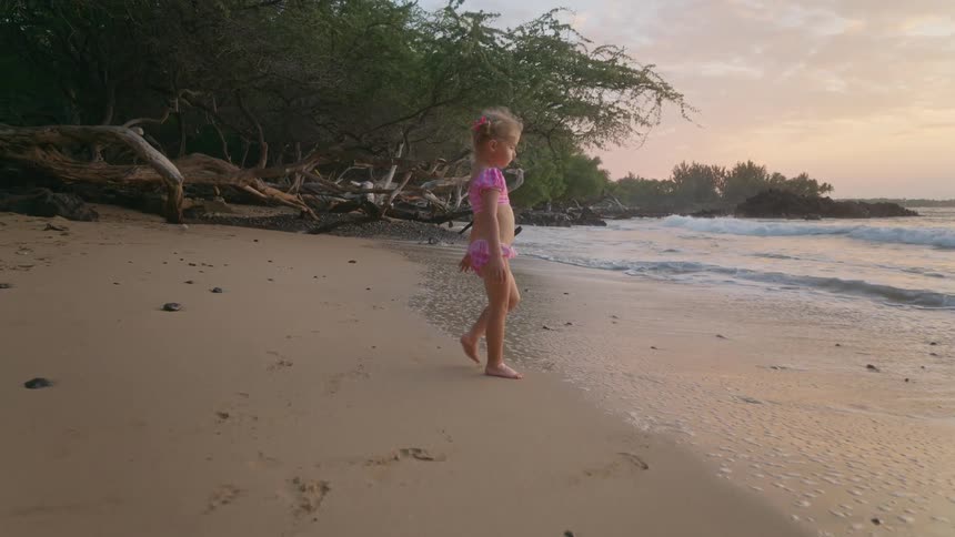 A young girl in a pink swimsuit playing on the beach at sunset stepping out of the water and doing acrobatic moves on the sand as the warm light glows behind her