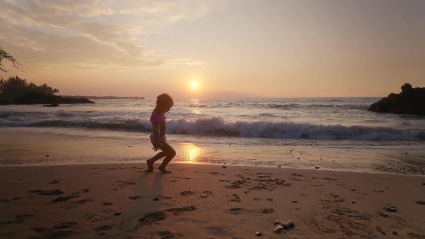 Child playing on the beach at sunset running toward the waves squatting to pick up a rock then dashing away before the water reaches them