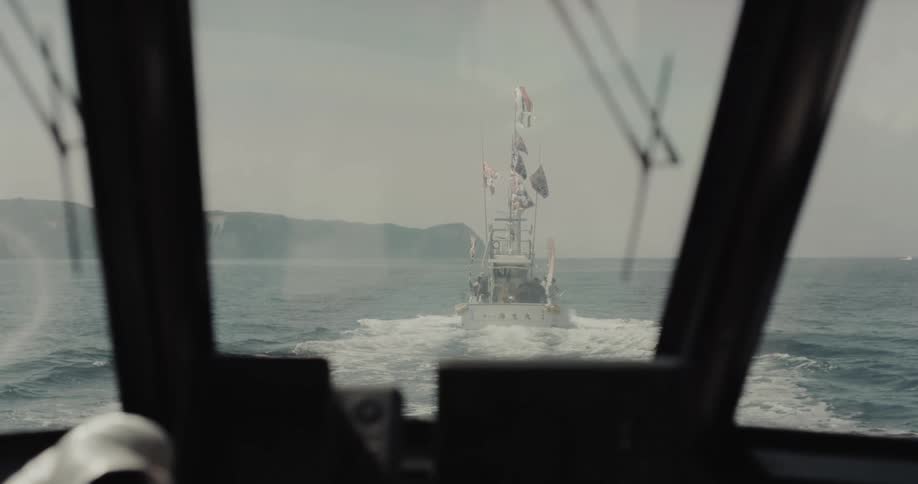 A boat trailing behind another across the ocean on a cloudy day the water choppy and the sky muted as they move forward