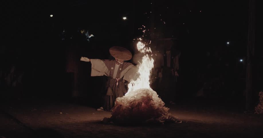 Person wearing traditional clothing performing a ritual beside a bonfire in an outdoor nighttime setting
