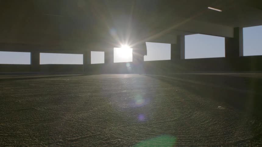A silhouetted figure in tall boots walks across an empty parking garage their form backlit by a brilliant sun flaring through an open window