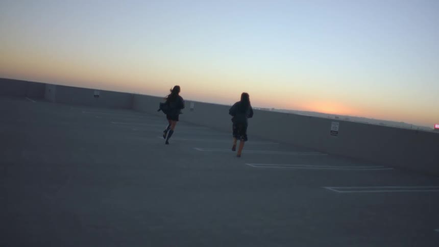 At dusk two people run across an empty rooftop parking lot toward the horizon their jackets and hair streaming in the wind as the vast concrete expanse and gradient sky