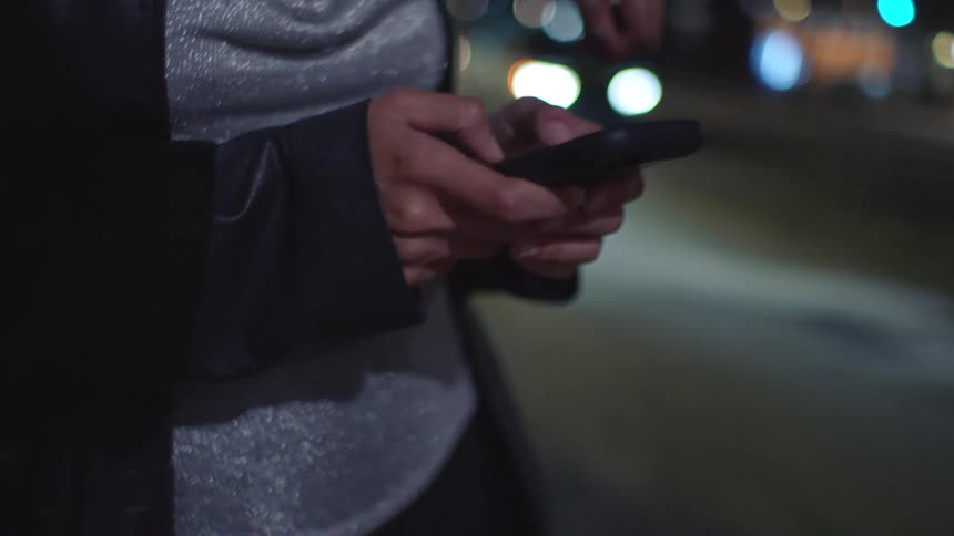 In a nighttime urban setting two women stand together with one wearing a silver top and a black leather jacket this woman is shown in a closeup shot as she uses both thumbs to type on a smartphone