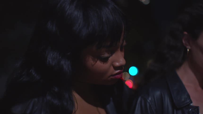 Two women in black leather jackets the woman in the foreground with long dark hair looking downward in soft side light while colorful city bokeh