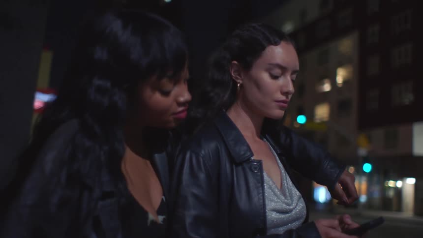 Two women in a nighttime urban setting one resting an arm on the other while looking at a smartphone