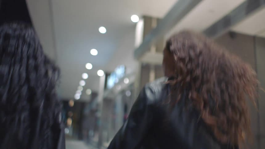 Two young black women wearing black jackets walking side by side along a nighttime sidewalk moving quickly as the streetlights cast long reflections across the pavement