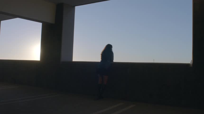 A young woman standing on a balcony at sunset gazing out at the sky at sunset