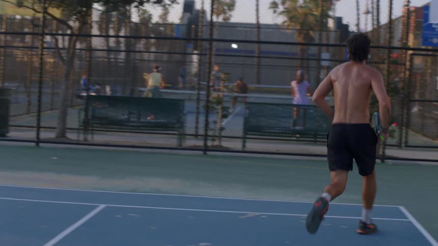 Shirtless man wearing glasses playing tennis on an outdoor court at midday