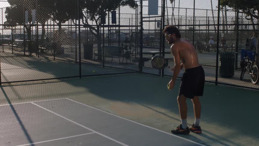 A shirtless man with sunglasses and tattoos hits a yellow tennis ball with a paddle on an outdoor court while another player walks in the background