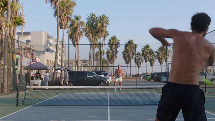 From a dynamic position behind one player two shirtless men rally on a beachside pickleball court surrounded by palm trees and the bustle of a sunny afternoon