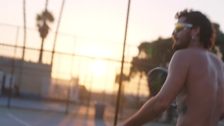A shirtless tattooed man serves a ball with a paddle on a sunny outdoor court