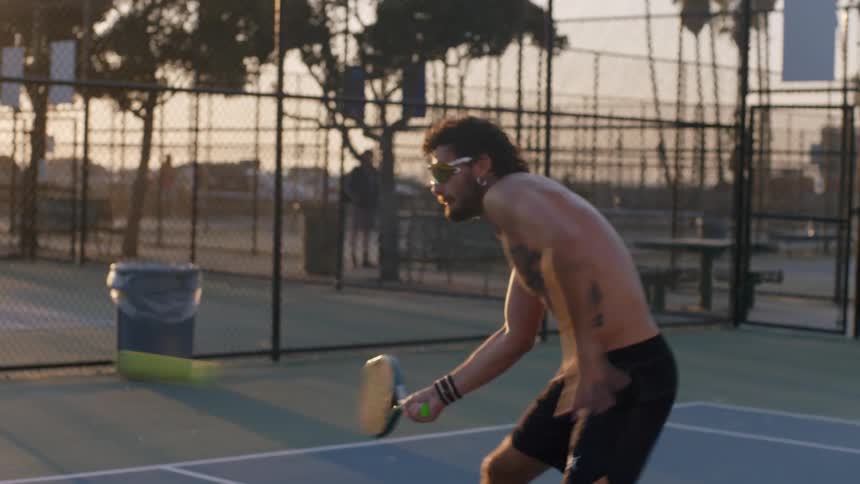 A shirtless tattooed man wearing sunglasses and black shorts moves across a blue pickleball court swinging his paddle to return shots during a game played outdoors at sunset