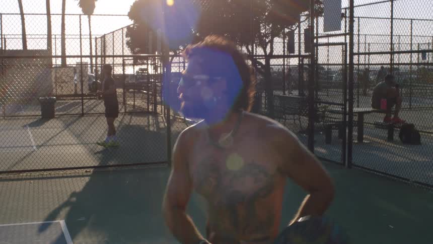 A shirtless man wearing sunglasses and a chain is playing pickleball on an outdoor court with another player visible in the background