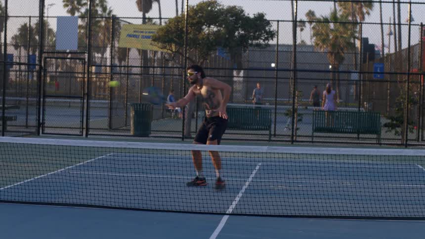 A shirtless man returns a tennis serve with a powerful swing sending the ball back to his opponent across the net