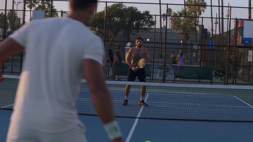 Two young men playing tennis at sunset one shirtless in black shorts and the other in a white tshirt and white shorts