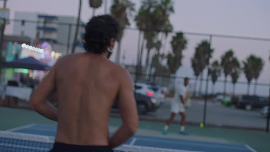 Two young men playing tennis on a court at sunset one shirtless wearing earrings and a necklace the other in a white tshirt and white trunks