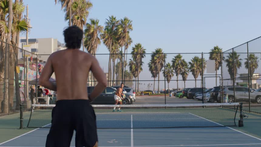 A shirtless man in shorts is playing a paddle sport hitting a ball across the net to an opponent on an outdoor court lined with palm trees and a parking lot behind a fence