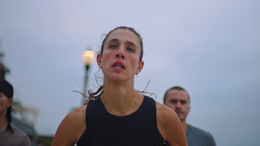 The face of a woman staring straight ahead while running with two people running behind her