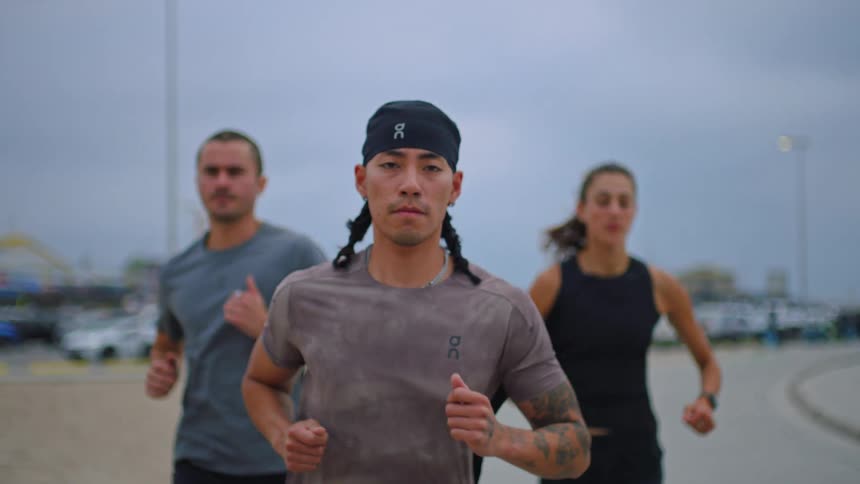 Three runners a man in a bandana with pigtails in the foreground and a man and woman slightly out of focus behind him are jogging outdoors on an overcast day