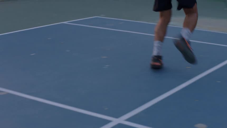 Male tennis player on a court at midday wearing black sneakers and white socks as he moves into position to hit the ball