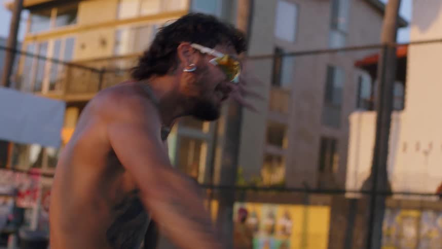 In warm goldenhour light on an urban outdoor court a shirtless tattooed man wearing reflective wraparound sunglasses and layered necklaces grips a round perforated paddle midplay with a chainlink fence and apartment buildings