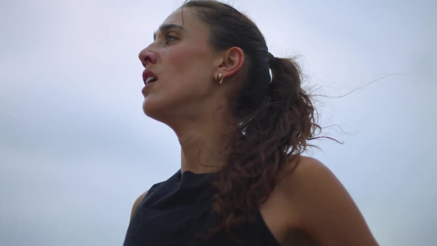 A young woman with a focused slightly exhausted expression gazes upward against an overcast sky her curly hair pulled back in a ponytail