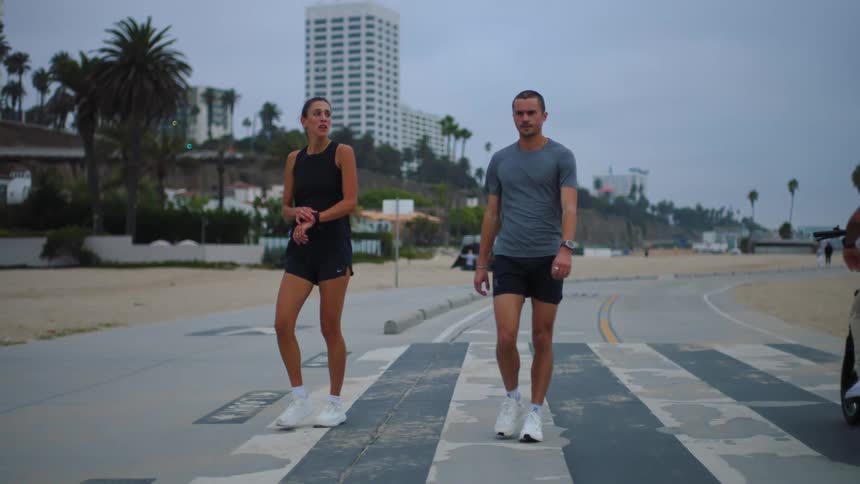 Two people walk side by side across a pedestrian crosswalk on a road near the beach with buildings in the background