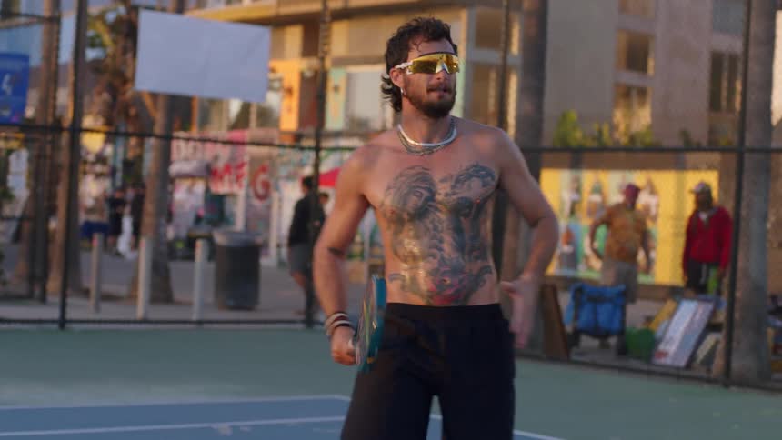 A shirtless heavily tattooed player in mirrored wraparound shades and layered chains strides midswing with a perforated paddle while chainlink fencing street art and a few onlookers blur in the background