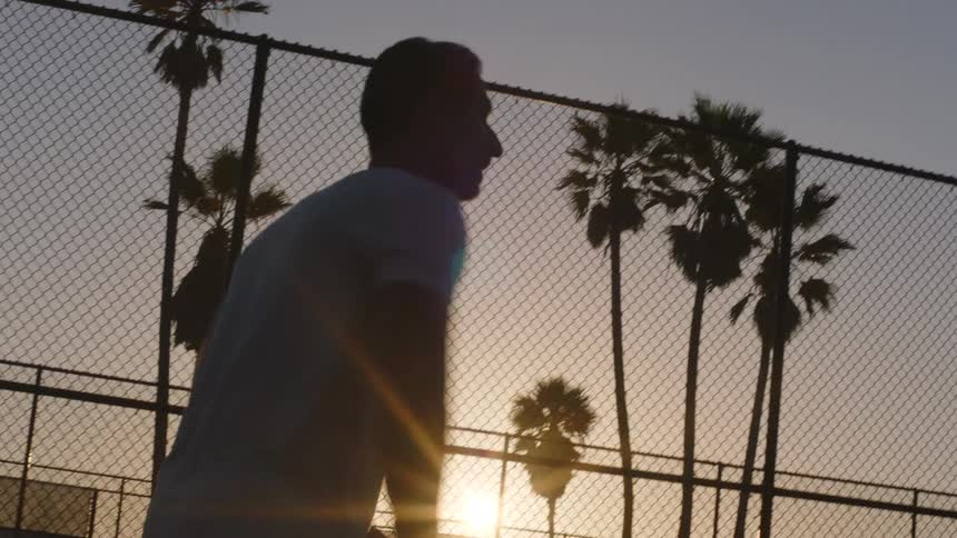 At sunset a silhouetted man in a light tshirt is swinging a pickleball paddle near a chainlink fence around an outdoor court with palm trees