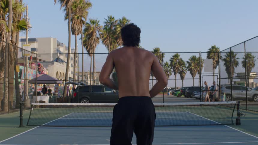 Two shirtless men play a game of pickleball on a sunny outdoor court surrounded by tall palm trees and parked cars the camera stays behind one player as he rallies the ball back and forth across the net with his opponent