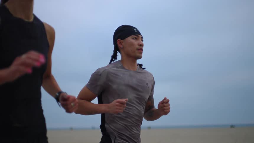 On an overcast beach a focused runner in a gray sweatdampened tshirt and a black headwrap with braids jogs in profile beside a partially visible companion arms pumping against a soft bluegray horizon