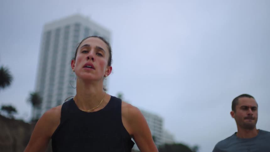 A woman in a black tank top and a man in a grey shirt run along a coastal path on an overcast day with tall palm trees and a white building in the background