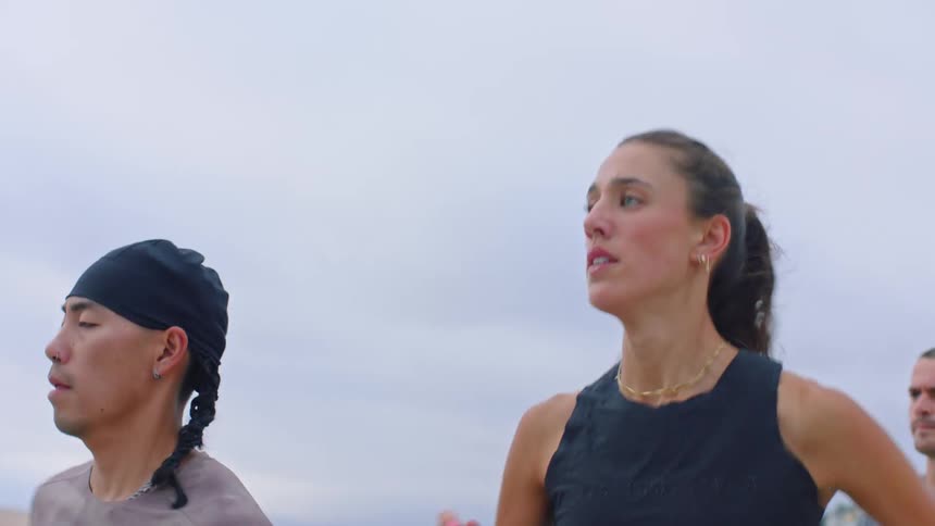 A group of people jogs along a sandy beach under an overcast sky with the camera focusing on a woman in a black tank top and a man in a headwrap as they run past coastal buildings in the background