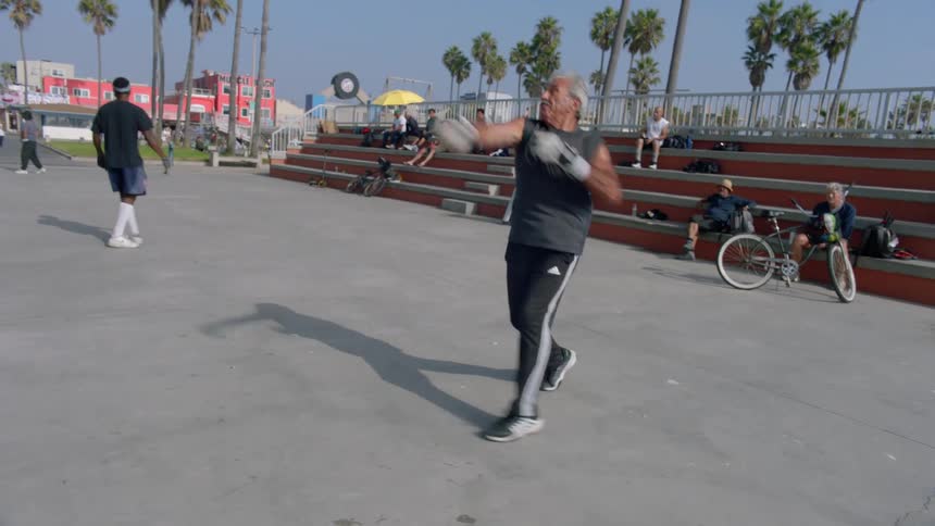An old man wearing a gray tank top is hitting a ball that is coming towards him using a glove
