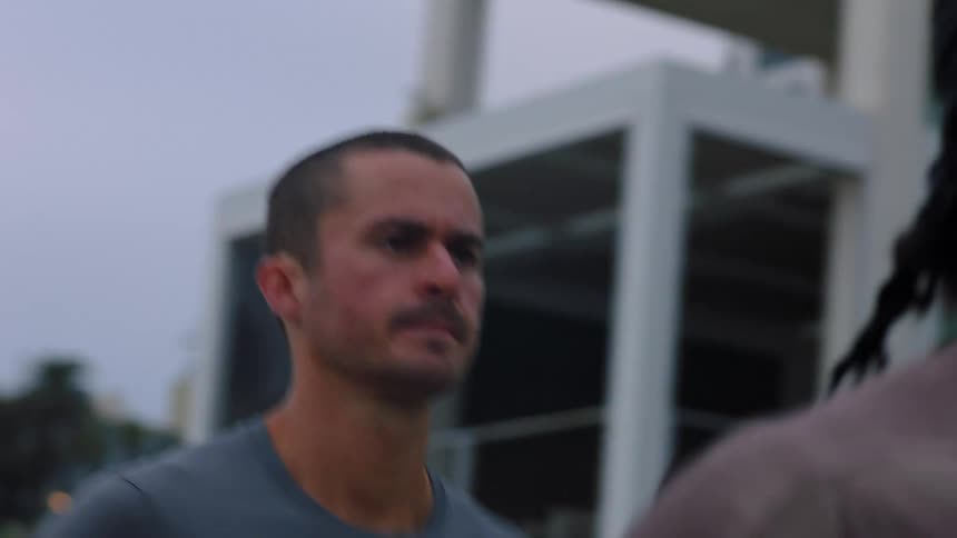 A man with a buzz cut and mustache maintains a steady determined expression while jogging outdoors past modern buildings during an athletic training session on a cloudy day with another man on the front
