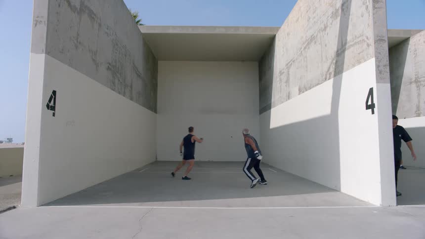 Four men play a game of handball in a large concrete outdoor court by hitting a ball against the front wall while moving across the pavement during a sunny day