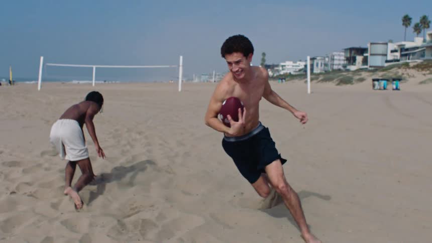 Young man runs across the beach carrying a football as another young man fails to tackle him