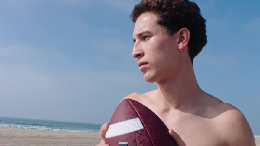 Young man stands on the beach holding a football